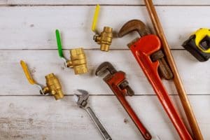 Plumbing tools including pipe wrenches, crescent wrench, copper pipe, tape measure, and brass ball valves on a wooden deck