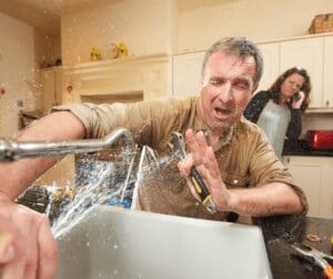 Man attempting DIY faucet repair with water spraying all over him while a woman, possibly his wife, calls a plumber in the background.