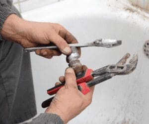 Man holding plumbing parts and wrenches while sitting in a bathtub, focusing on his hands.