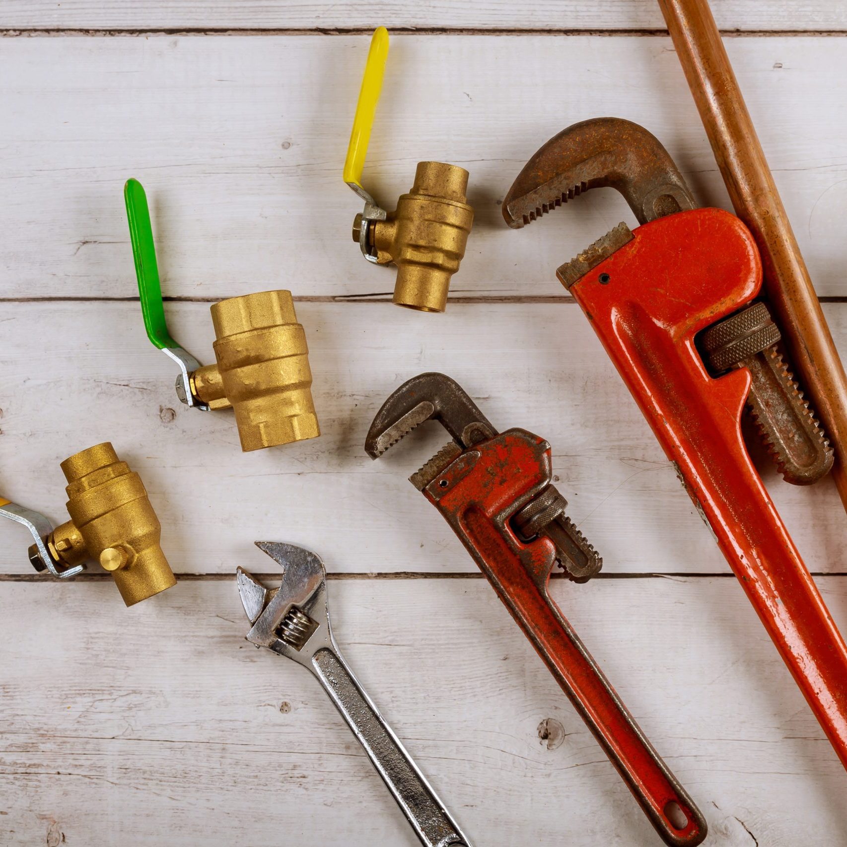 Plumbing tools including pipe wrenches, crescent wrench, copper pipe, tape measure, and brass ball valves on a wooden deck